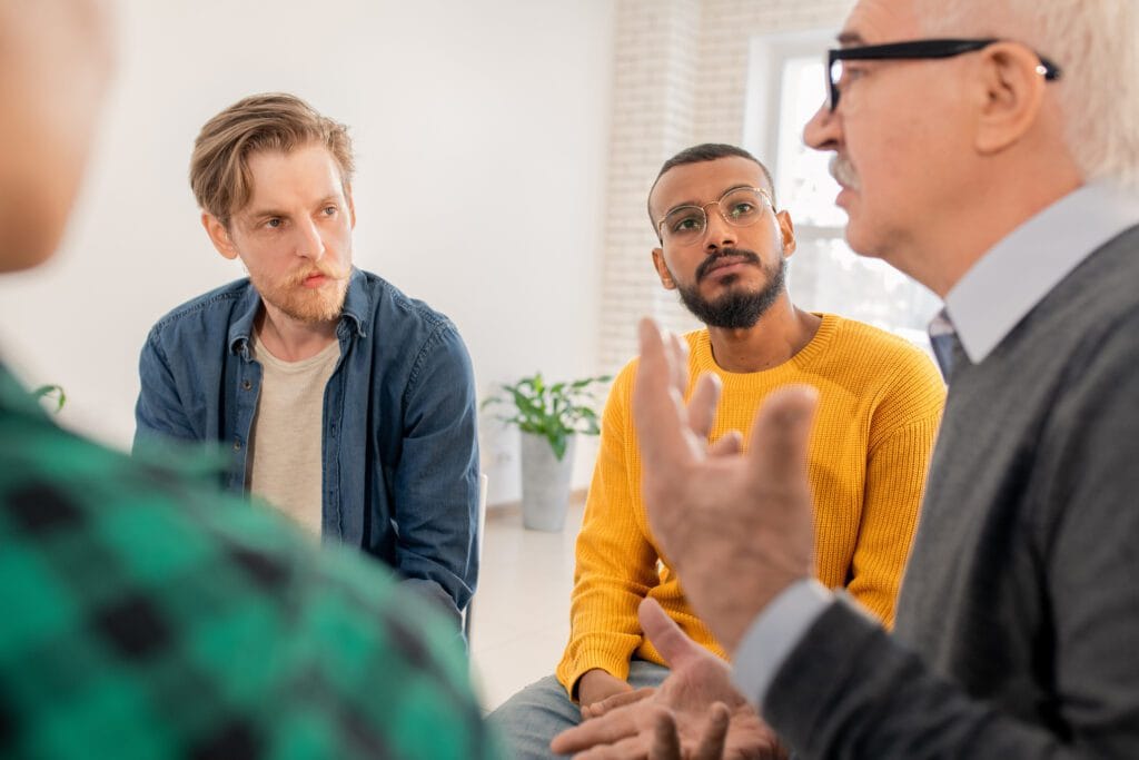 Two young men looking at senior groupmate talking about his problem at a session