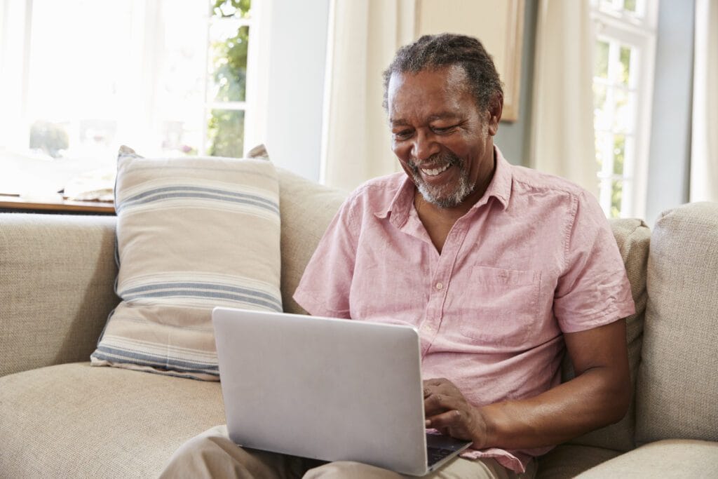 Senior Man Sitting On Sofa Using Laptop At Home Together
