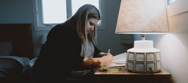 Blonde woman with rose forearm tattoo sitting at wooden desk with lamp in private bedroom writing in journal